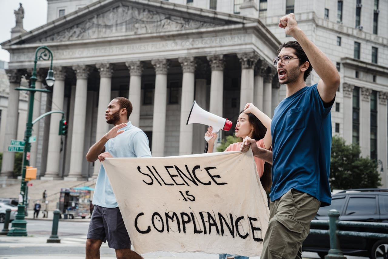 Demonstrators marching with a banner saying Silence is Compliance and a megaphone in front of a courthouse.