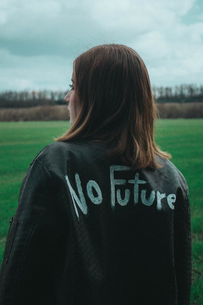 A woman with short hair wearing a jacket with No Future stands in an open field on an overcast day.